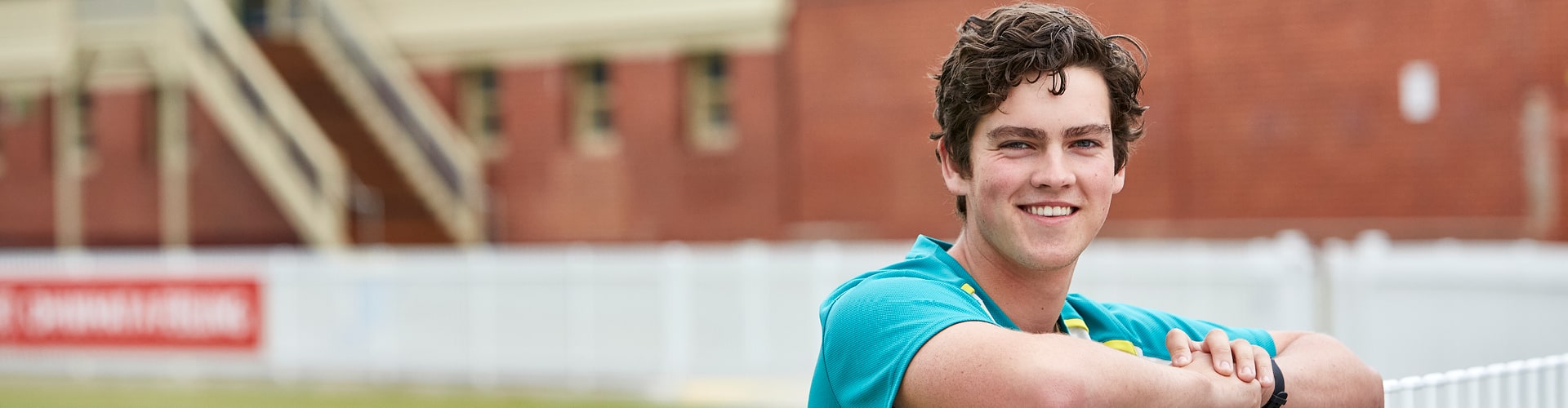 A Cricket Australia student resting on a fence at a cricket oval and smiling at the camera