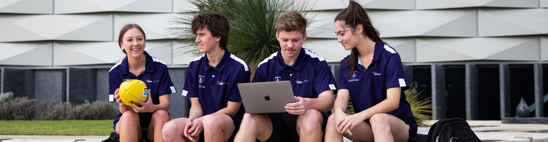 Four Fremantle Dockers students sitting outside, talking and working on a laptop.